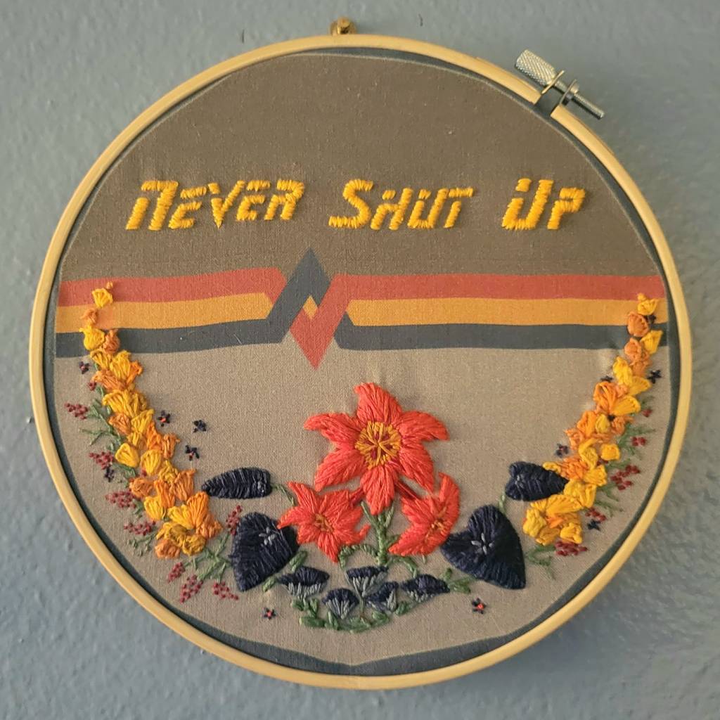 An embroidery hoop with a design similar to Wesley Crusher’s iconic pseudo-uniform sweater from Star Trek: TNG as the background fabric. Embroidered in the top/dark gray half is the words “Never Shut Up” embroidered in gold thread in the TNG title font. The Red/Yellow/Dark Blue bar and diamond design bisects the hoop. On the lower / light gray half are embroidered a garland of flowers from Star Trek scenes in matching gold, coral red, and dark blue.